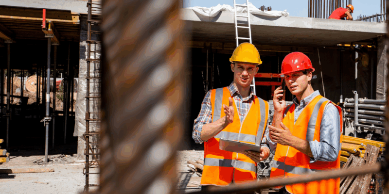 Two construction workers wearing safety vests and helmets discuss plans at a construction site. One holds a tablet, while the other gestures. Metal rebar is visible in the foreground, with a building and workers in the background.