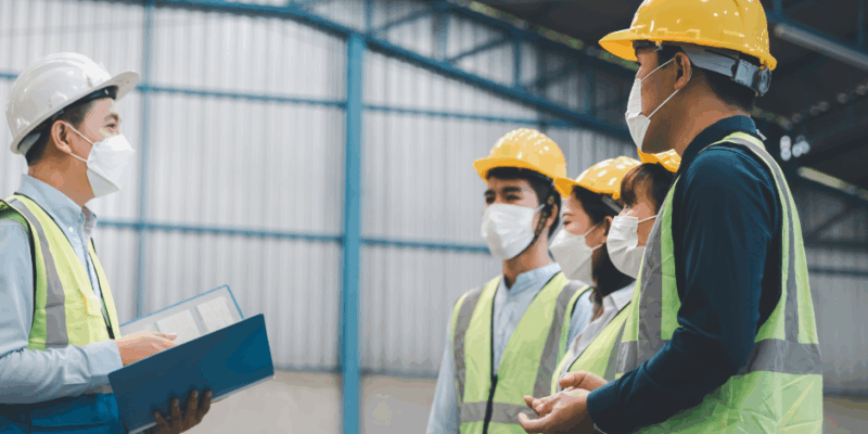 A group of workers wearing safety vests, hard hats, and face masks stands in a warehouse, listening to a supervisor holding a blue folder and giving instructions.