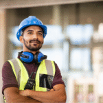 A construction worker wearing a blue hard hat, safety vest, and noise-canceling headphones stands with arms crossed, looking confident. A walkie-talkie is clipped to the vest, with a blurred indoor background.