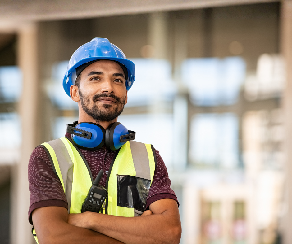 A construction worker wearing a blue hard hat, safety vest, and noise-canceling headphones stands with arms crossed, looking confident. A walkie-talkie is clipped to the vest, with a blurred indoor background.