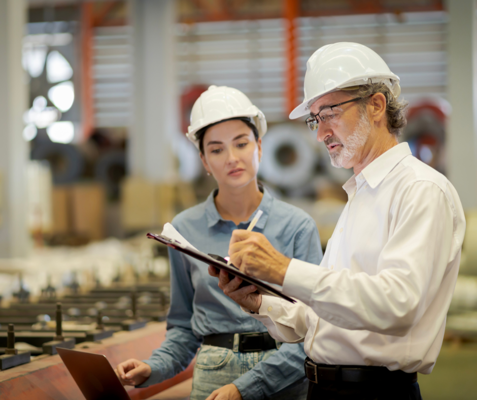 Two people wearing hard hats stand in a factory. One writes on a clipboard while the other watches attentively, holding a laptop. Industrial equipment and blurred machinery are visible in the background.