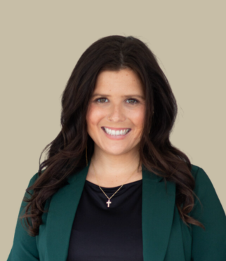 A woman with long dark hair, wearing a dark green blazer, black top, and a necklace with a cross pendant, smiling in front of a plain beige background.
