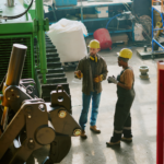 Two workers wearing yellow safety helmets and protective clothing stand in a factory, discussing machinery. Industrial equipment and supplies are visible in the background.