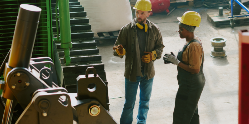 Two workers wearing yellow safety helmets and protective clothing stand in a factory, discussing machinery. Industrial equipment and supplies are visible in the background.