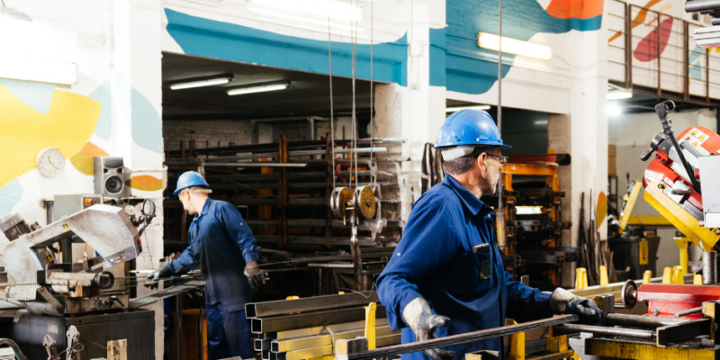 Two workers in blue uniforms and helmets operate machinery in a brightly lit metal workshop, surrounded by metal rods, tools, and colorful painted walls.