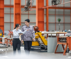 Two people, one in business attire and the other in work overalls, walk together in a large industrial warehouse, discussing documents amid machinery and metal equipment.