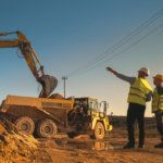 Two construction workers in safety vests and helmets observe an excavator loading dirt into a dump truck at a worksite during sunset. One worker gestures towards the truck, while power lines run overhead.
