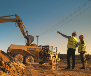 Two construction workers in safety vests and helmets observe an excavator loading dirt into a dump truck at a worksite during sunset. One worker gestures towards the truck, while power lines run overhead.