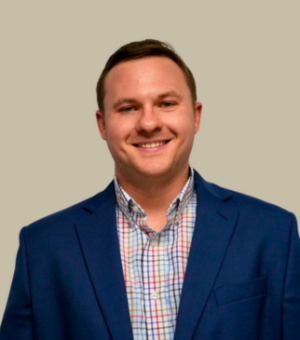 A man wearing a blue blazer over a multicolored checkered shirt is smiling at the camera against a plain, light beige background.