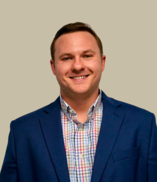 A man wearing a blue blazer over a multicolored checkered shirt is smiling at the camera against a plain, light beige background.
