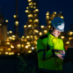 An engineer wearing a hard hat and high-visibility jacket works on a laptop at night in front of a brightly lit industrial plant.