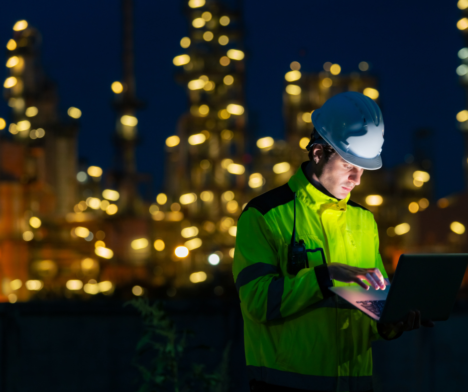 An engineer wearing a hard hat and high-visibility jacket works on a laptop at night in front of a brightly lit industrial plant.