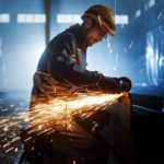 A worker wearing safety gear and a helmet uses an angle grinder on metal, producing bright sparks in a dimly lit industrial setting.