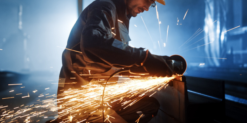 A worker wearing safety gear and a helmet uses an angle grinder on metal, producing bright sparks in a dimly lit industrial setting.