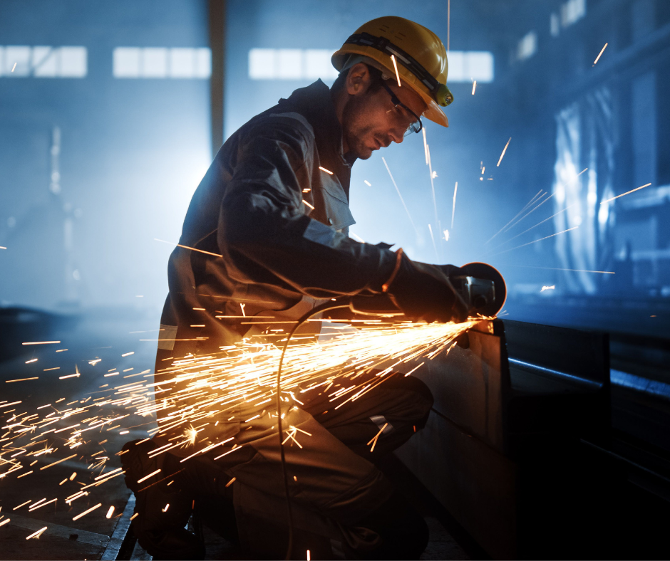 A worker wearing safety gear and a helmet uses an angle grinder on metal, producing bright sparks in a dimly lit industrial setting.