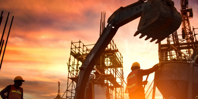 Construction workers wearing safety gear operate heavy machinery and work on scaffolding at a construction site during sunset, with a dramatic sky of orange and yellow hues in the background.